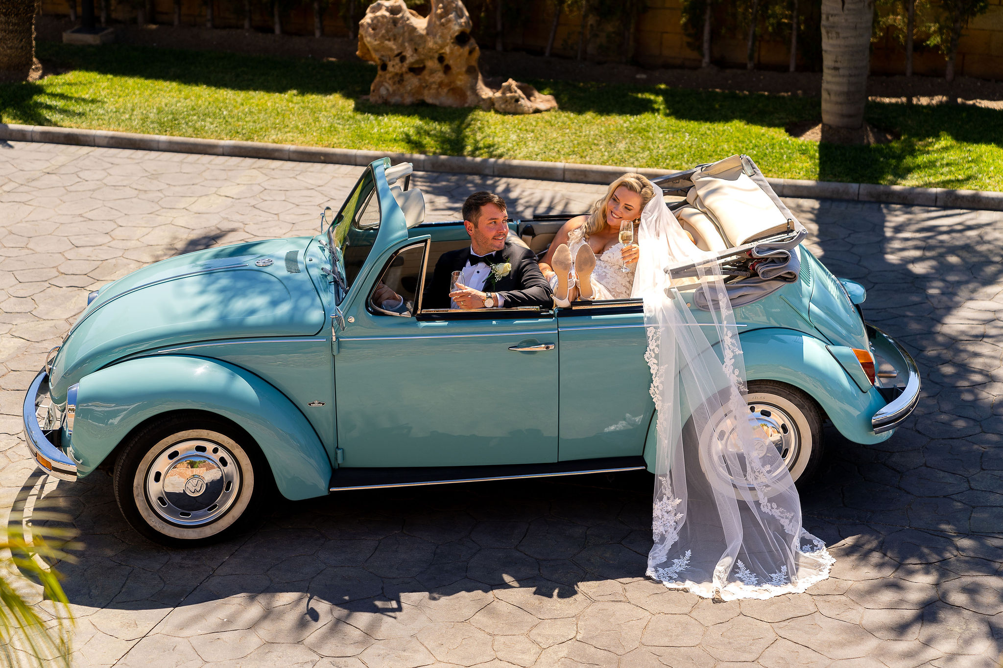 The bride, wearing a white wedding dress and veil, is seen sipping champagne sitting in the back seat of a classic Volkswagen Beetle. The groom, attired in black tie is also celebrating drinking champagne in the front seat. One lucky couple will win a wedding competition.