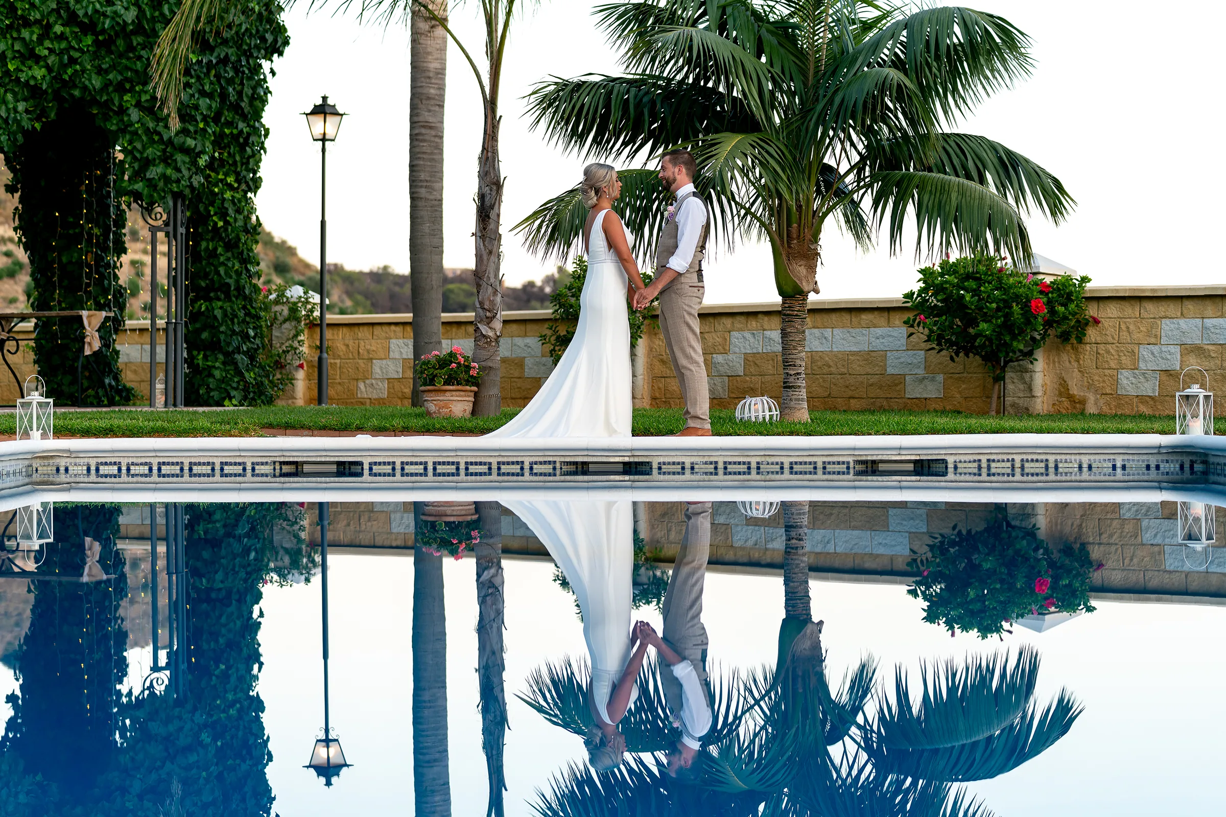 RG02-536 – Rachel Smyth and George McMahon Wedding, Nerja, Spain Bride and groom holding hands beside a swimming pool beneath palm trees, reflected in the water at a wedding in Nerja, Spain.
