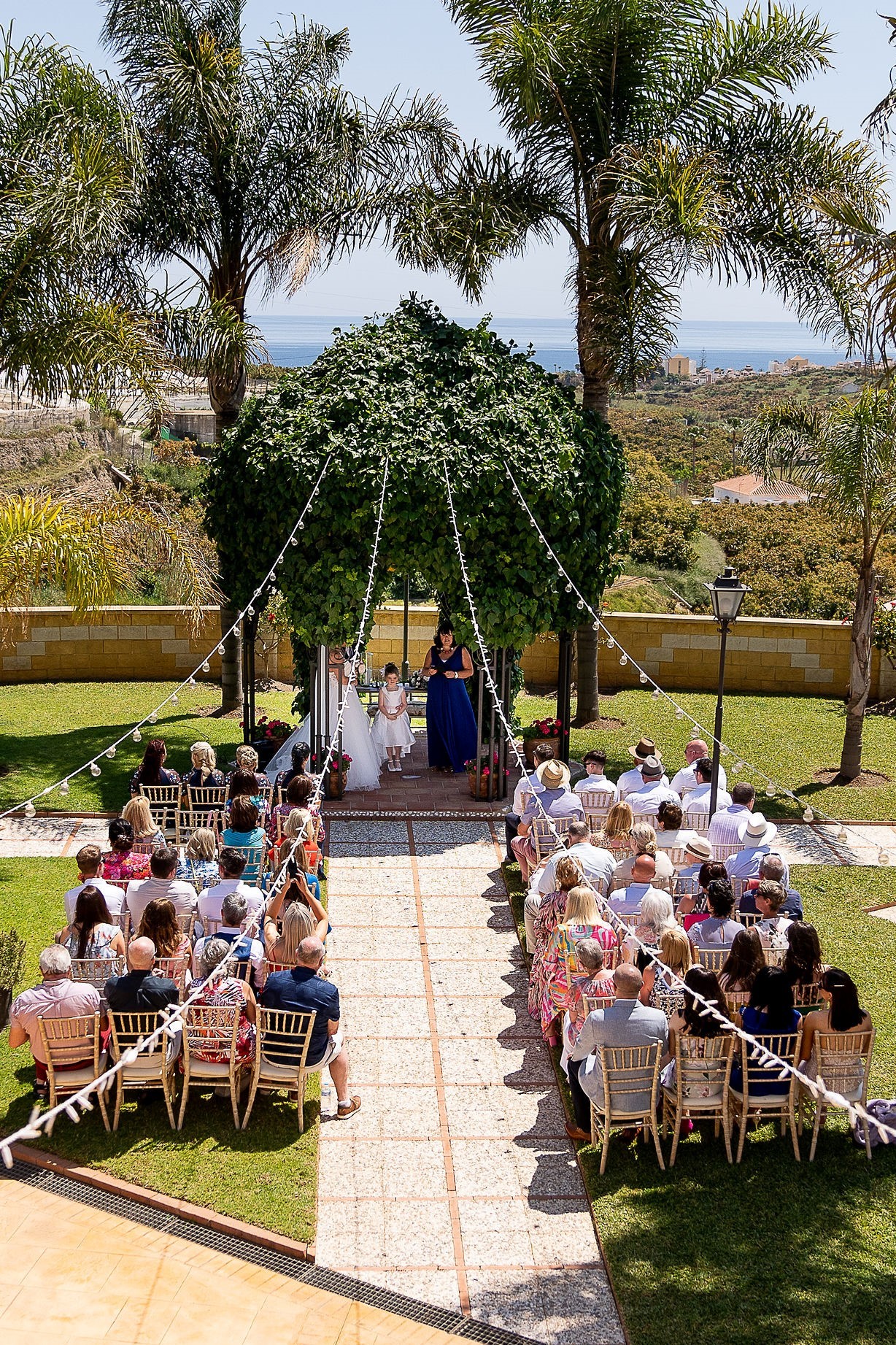 Outdoor wedding ceremony at Cortijo Maria Luisa in Spain with guests seated under palm trees and a sea view in the background – photo by Dougie Farrelly, Silverscreen.ie.