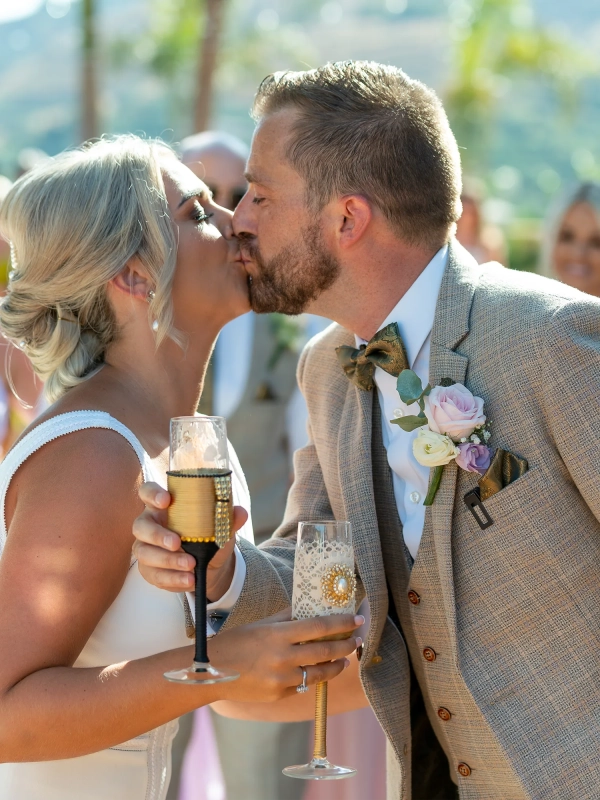 Rachel Smyth and George McMahon kissing while holding champagne glasses during their wedding celebrations in Nerja, Spain.