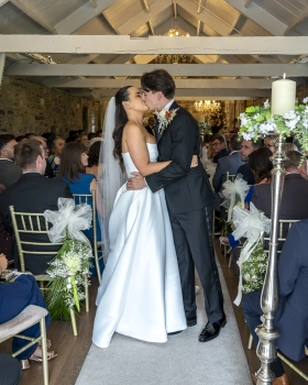 Newlywed couple kissing during their wedding ceremony at Ballymagarvey Village, surrounded by seated guests