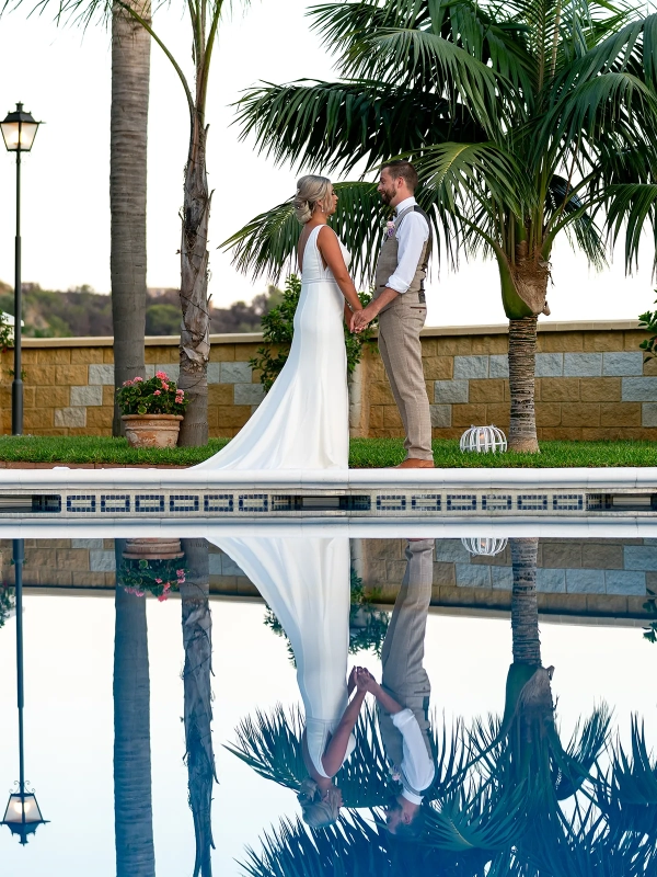 Bride and groom holding hands beside a swimming pool beneath palm trees, reflected in the water at a wedding in Nerja, Spain.