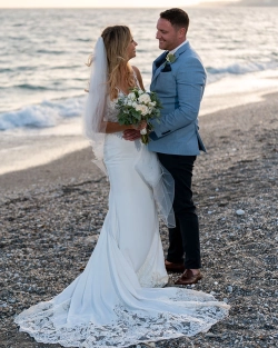 A beautiful moment by the Mediterranean Sea as Ruth and James enjoy their wedding celebrations in Nerja. Photo: Dougie Farrelly / Silverscreen Productions (Photography & Video). Ruth and James smiling at each other on a beach in Nerja, with the bride holding a white floral bouquet and wearing a lace wedding dress with a flowing train.