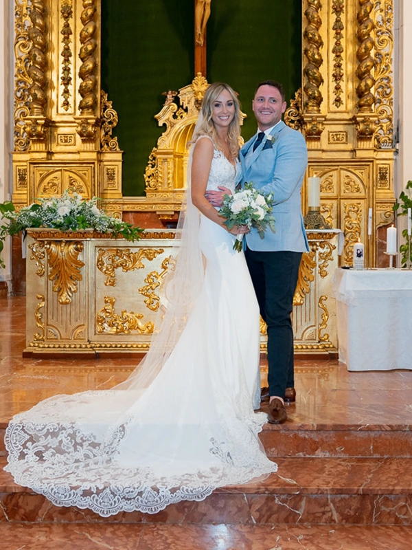 Ruth and James standing together in front of an ornate golden church altar, with the bride wearing a lace wedding dress and holding a white floral bouquet.