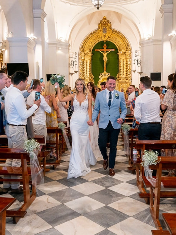 Ruth and James walking hand in hand down the church aisle after their wedding ceremony, smiling as guests celebrate on either side.