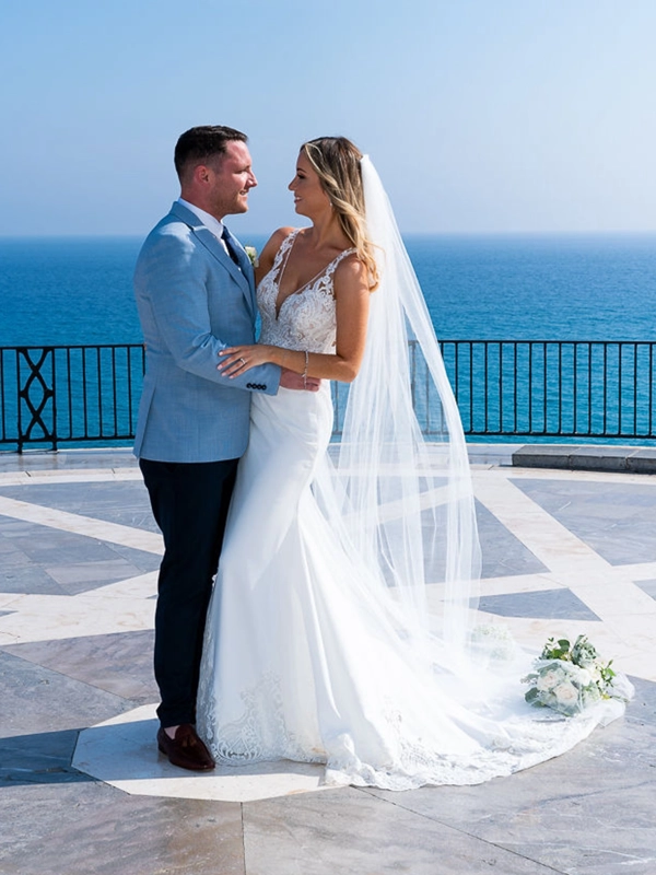 Ruth and James embracing on a terrace overlooking the Mediterranean Sea, with the bride's long veil and lace wedding dress train flowing behind her.