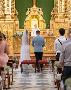 Ruth and James exchange vows before an ornate golden altar during their church wedding ceremony in Spain. Photo: Dougie Farrelly / Silverscreen Productions (Photography & Video). Ruth and James standing before a priest at an ornate golden church altar during their wedding ceremony, viewed from behind.