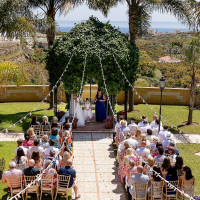 A breathtaking outdoor ceremony under the Andalusian sun at Cortijo Maria Luisa, beautifully captured by Irish wedding photographer Dougie Farrelly of Silverscreen.ie. © Dougie Farrelly – Silverscreen.ie. All rights reserved. Outdoor wedding ceremony at Cortijo Maria Luisa in Spain with guests seated under palm trees and a sea view in the background – photo by Dougie Farrelly, Silverscreen.ie.